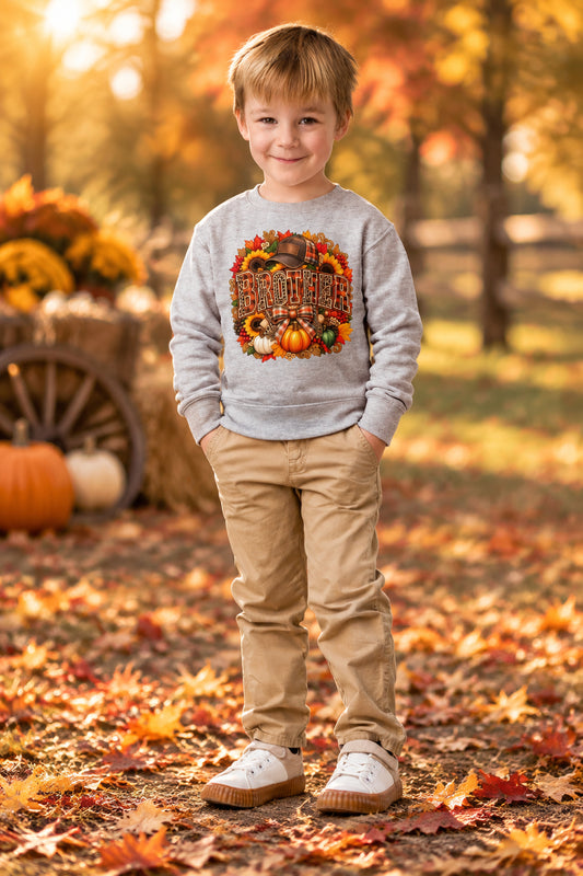 A young boy wearing a light gray crewneck sweatshirt with a colorful 'Thanksgiving' graphic featuring pumpkins and autumn leaves, paired with khaki pants and white slip-on shoes, standing against a plain white background.