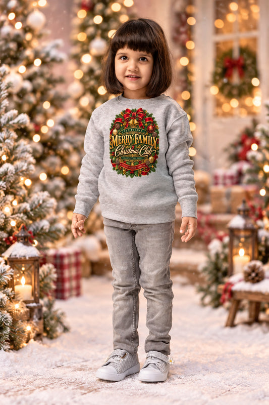 A young girl with dark hair stands against a white background wearing a light gray sweatshirt with a colorful 'Merry Family Christmas' graphic, gray pants, and gray sneakers.