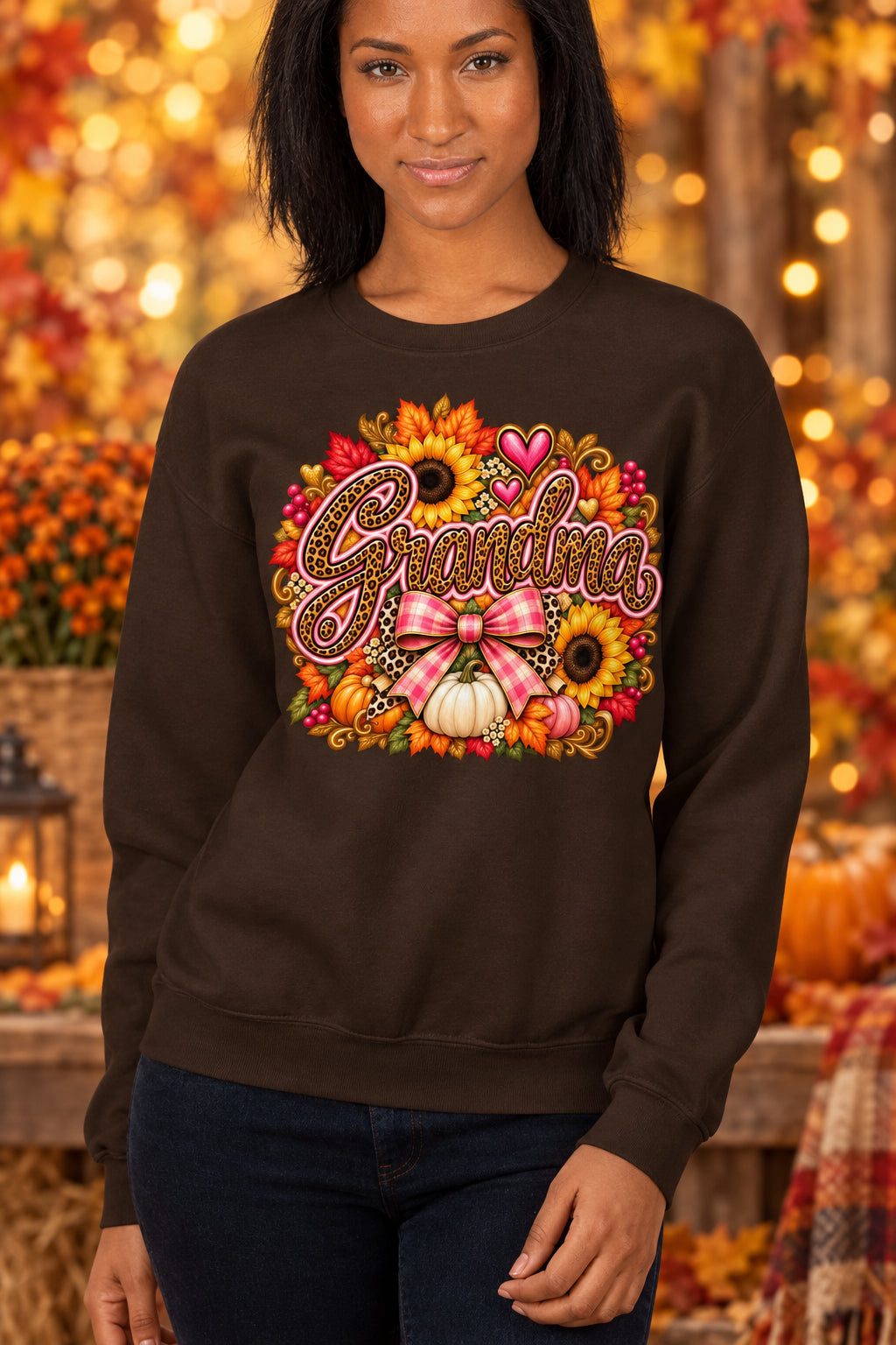 A woman wearing a dark brown crewneck sweatshirt with a colorful autumn-themed graphic that reads 'Grandma' in script, surrounded by sunflowers, pumpkins, hearts, and a pink bow.