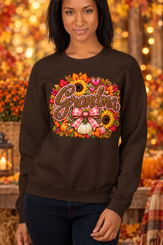 A woman wearing a dark brown crewneck sweatshirt with a colorful autumn-themed graphic that reads 'Grandma' in script, surrounded by sunflowers, pumpkins, hearts, and a pink bow.