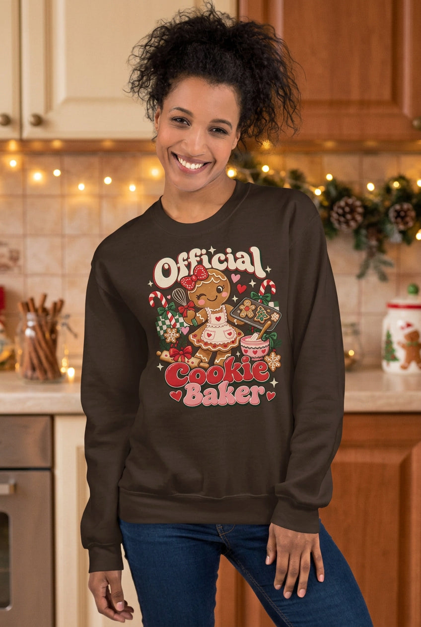 A woman wearing a dark brown crewneck sweatshirt with a colorful graphic that reads 'Official Cookie Baker' featuring a gingerbread man and baking theme, paired with blue jeans against a two-tone studio background.