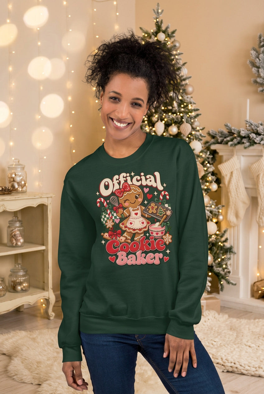 A woman wearing a dark green crewneck sweatshirt with a colorful graphic that reads 'Official Cookie Baker' featuring a gingerbread cookie illustration, posed against a two-tone gray and beige background.