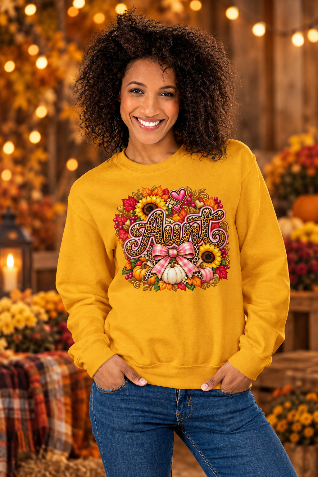 A smiling woman with curly hair wears a mustard yellow sweatshirt featuring a colorful autumn graphic that reads 'Aunt' surrounded by pumpkins, sunflowers, and hearts. She is posing in front of a split gray and white textured wall.