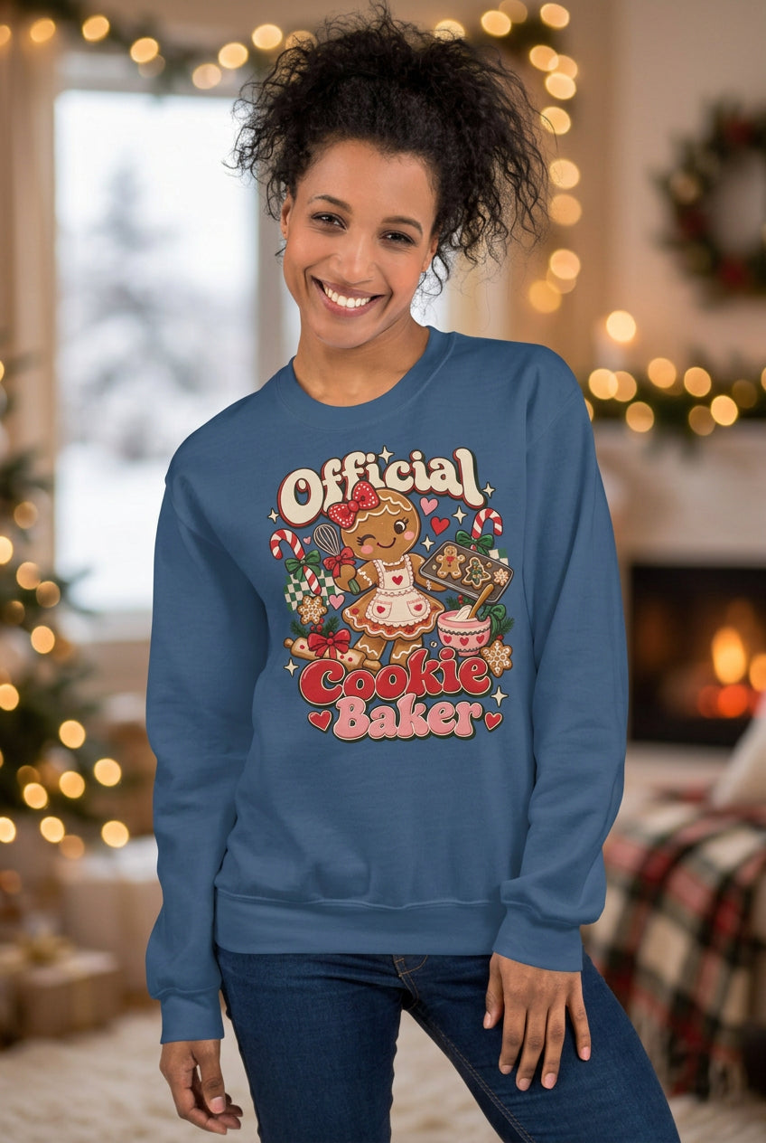 A woman wearing a slate blue crewneck sweatshirt with a colorful graphic that reads 'Official Cookie Baker' featuring a cartoon character, posed against a two-tone gray and beige background.