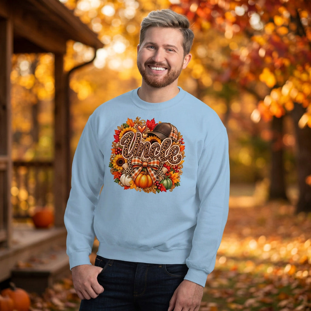 A smiling man wearing a light blue crewneck sweatshirt with a colorful autumn-themed graphic that reads 'Uncle' surrounded by pumpkins, leaves, and a plaid bow. He is standing against a white painted brick wall.
