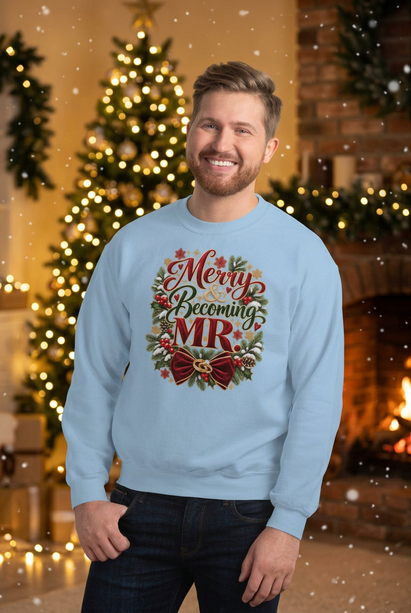 A smiling man wearing a light blue crewneck sweatshirt with a 'Merry Becoming MR' holiday wreath graphic, standing against a white brick wall.