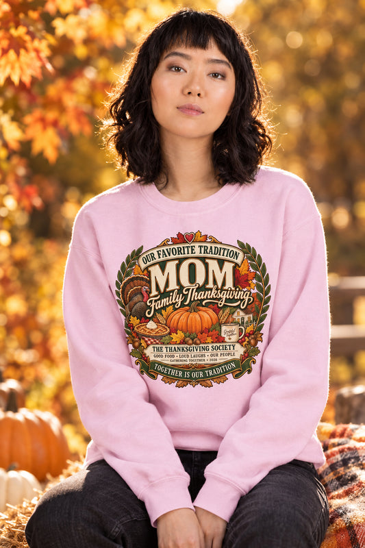 A woman wearing a light pink crewneck sweatshirt with a large, detailed autumn-themed graphic on the chest that reads 'Our Favorite Tradition Mom Family Thanksgiving' with pumpkins, turkeys, and fall leaves.