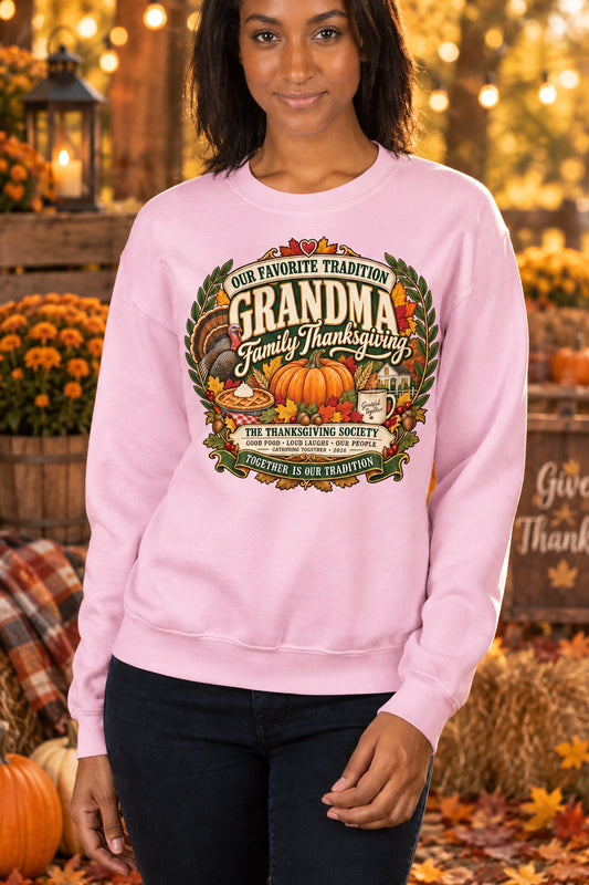 A woman wearing a light pink crewneck sweatshirt with a large, detailed Thanksgiving graphic on the chest. The graphic features a pumpkin, autumn leaves, and the text 'Our Favorite Tradition GRANDMA Family Thanksgiving'.