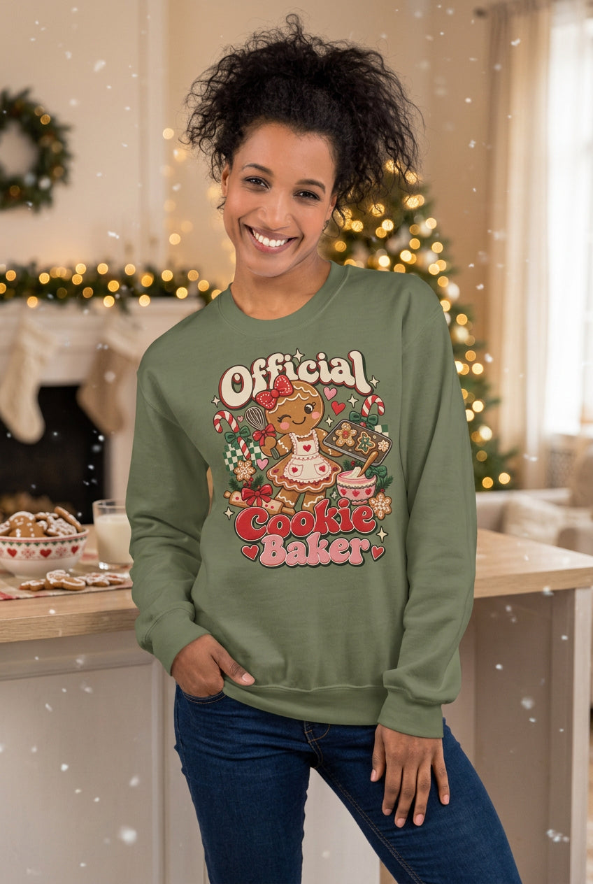 A woman wearing an olive green crewneck sweatshirt with a colorful graphic that reads 'Official Cookie Baker' featuring a cartoon character and desserts, paired with blue jeans, set against a two-tone gray and beige background.