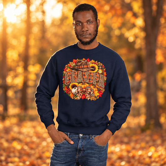 A man wearing a navy blue crewneck sweatshirt with a large, colorful autumn-themed graphic on the chest that reads 'GRANDPA' surrounded by fall leaves, pumpkins, and a cowboy hat. He is also wearing blue jeans, set against a plain white background.