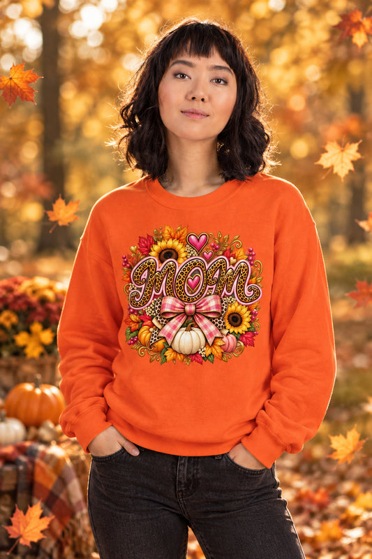 A woman wearing a bright orange crewneck sweatshirt with a colorful autumn-themed graphic on the chest featuring the word 'MOM' in brown glitter-style lettering surrounded by sunflowers, pumpkins, hearts, and a pink bow.