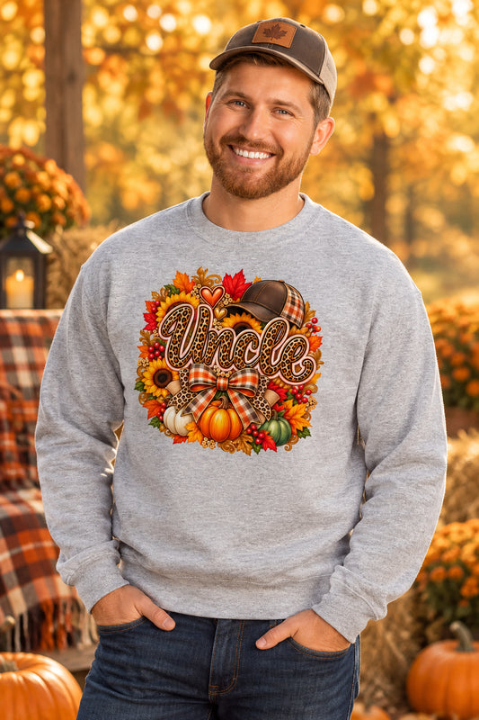 A smiling man wearing a light gray crewneck sweatshirt with a colorful autumn-themed graphic that reads 'Uncle' surrounded by pumpkins, leaves, and a plaid bow. He is standing against a white brick wall.