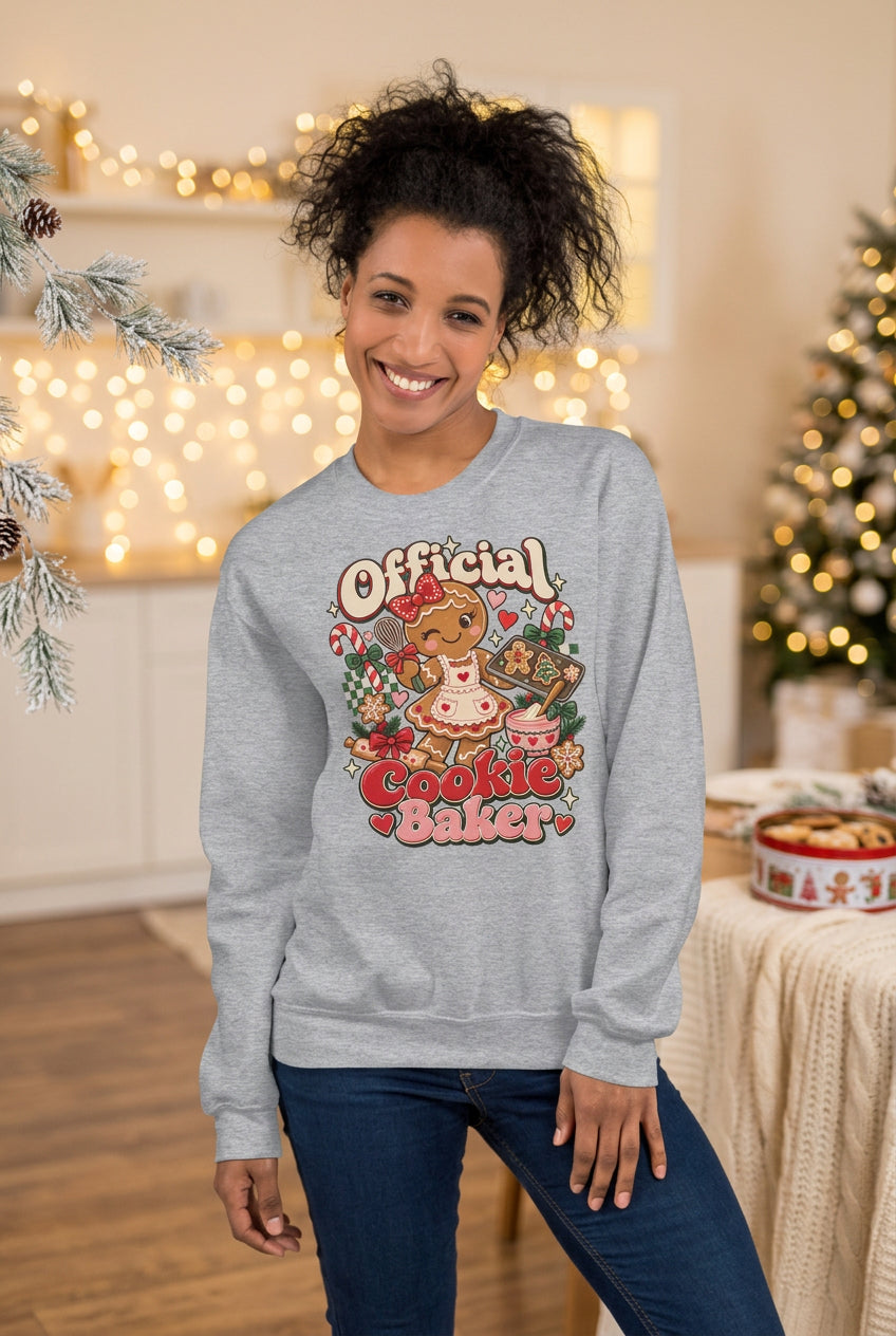 A woman wearing a light gray crewneck sweatshirt with a colorful retro graphic that reads 'Official Cookie Baker' featuring a gingerbread cookie and holiday treats, paired with dark blue jeans, set against a split gray and beige background.
