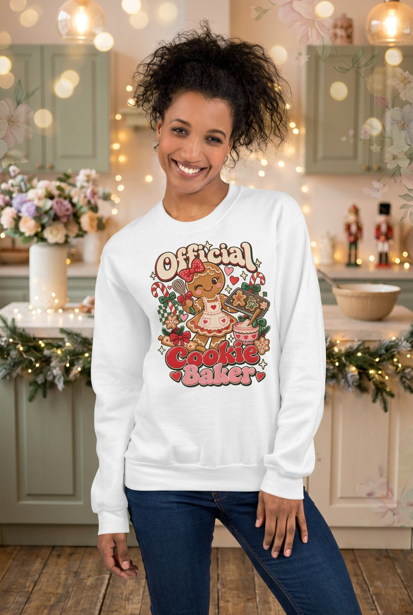 A woman wearing a white crewneck sweatshirt with a colorful graphic that reads 'Official Cookie Baker' featuring a gingerbread cookie and a pink cupcake, posed against a split gray and beige background.