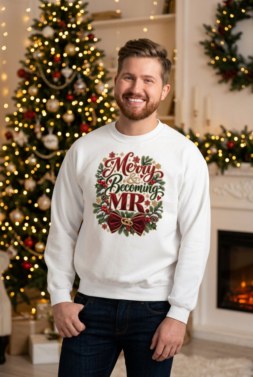 A smiling man wearing a white crewneck sweatshirt with a 'Merry Becoming MR' Christmas wreath graphic, standing against a white brick wall.