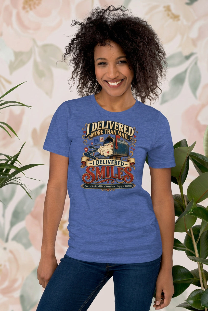 A smiling woman with curly dark hair stands indoors wearing a heathered blue graphic T-shirt and dark jeans, with large green houseplants on both sides against a light wall.