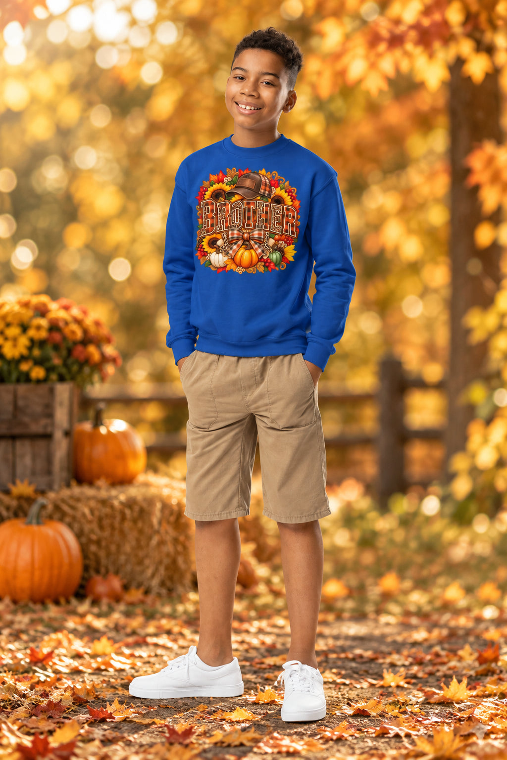 A young boy stands against a plain white background wearing a royal blue crewneck sweatshirt with a large colorful autumn graphic on the chest, khaki shorts, and white sneakers.